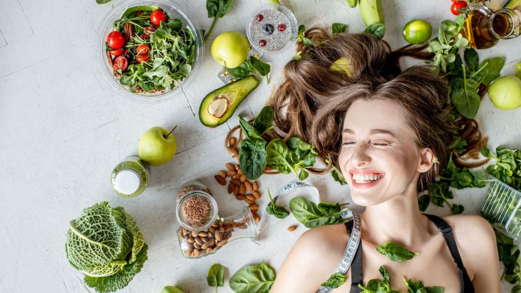 A woman smiles while lying on a white surface surrounded by fruits, vegetables, nuts, and leafy greens.