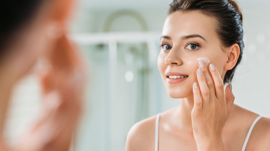 A woman applies cream to her cheek while looking in a mirror.