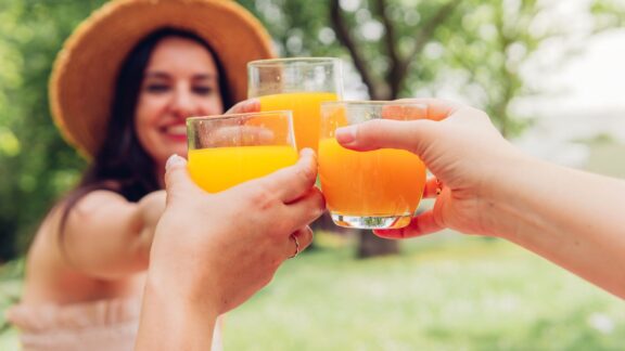Three people clinking glasses of orange juice outdoors, with a smiling woman in the background wearing a straw hat.