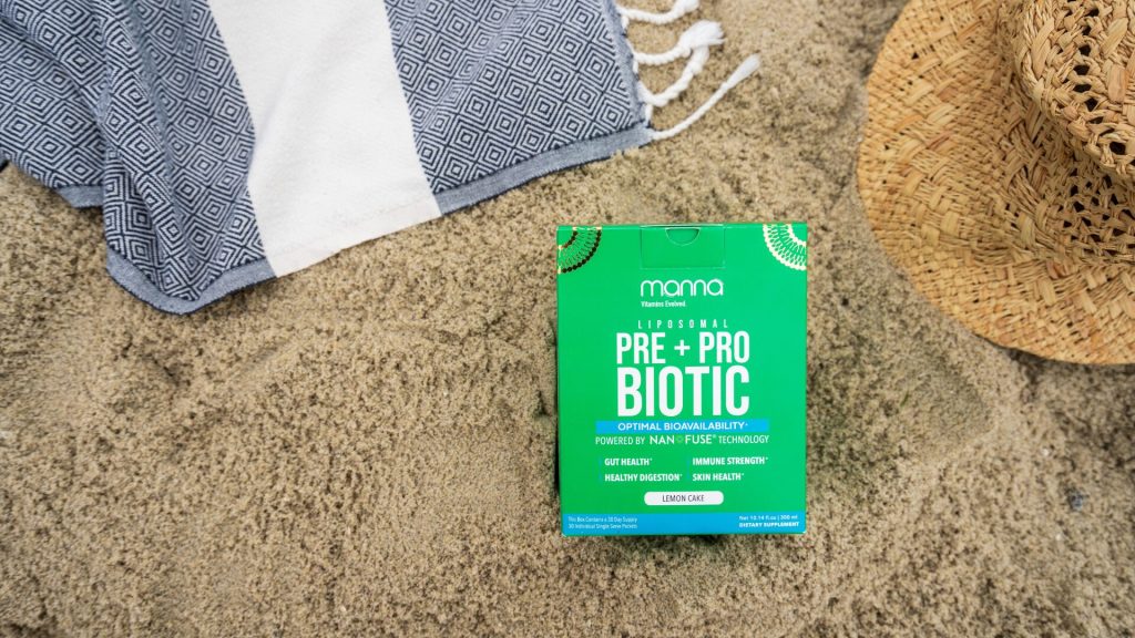 A green box of prebiotic and probiotic supplements rests on sandy ground with a striped towel and woven hat nearby.
