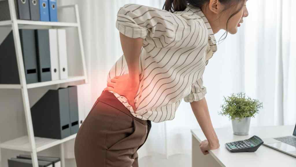 Woman in striped shirt and brown pants holds her lower back in pain, standing by a desk with a calculator and a plant.