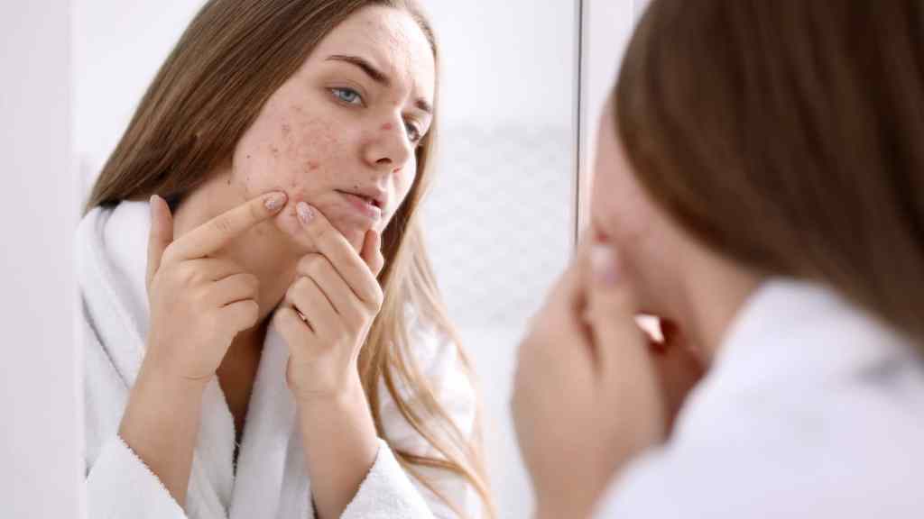 A woman in a white robe examines acne on her face while looking in a mirror.