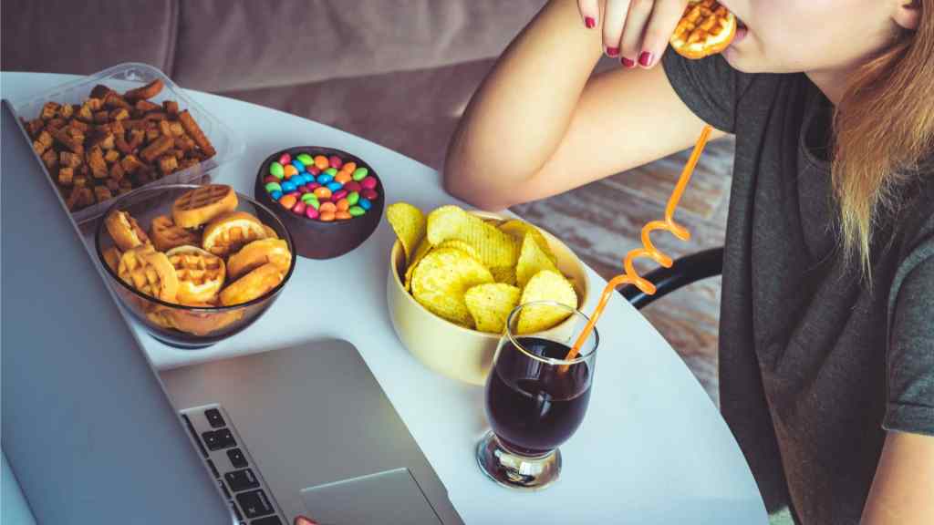 Person eating snacks at a table with a laptop. Bowls contain chips, waffles, colorful candies, and a drink with a curly straw.