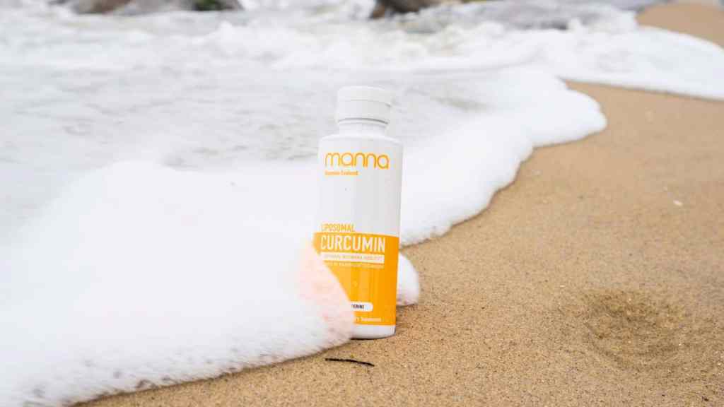 A bottle of Momo Liposomal Curcumin supplement is placed upright on a sandy beach near incoming ocean waves.