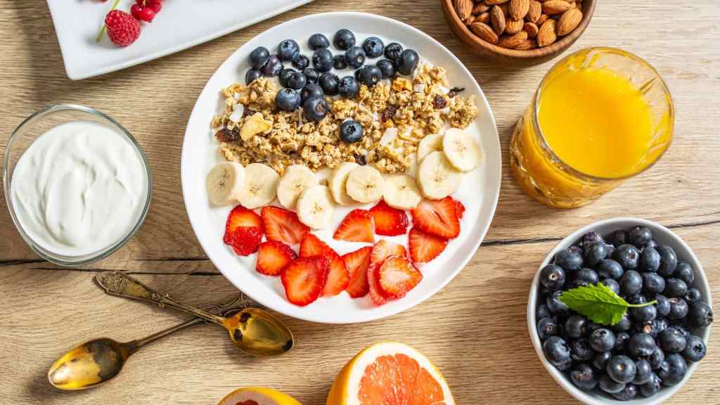A breakfast spread includes yogurt, granola with bananas, strawberries, blueberries, almonds, orange juice, and a halved grapefruit on a wooden table.