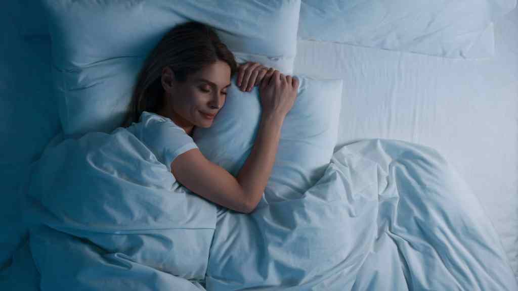 A person is sleeping peacefully in a bed with white sheets, wearing a white shirt.