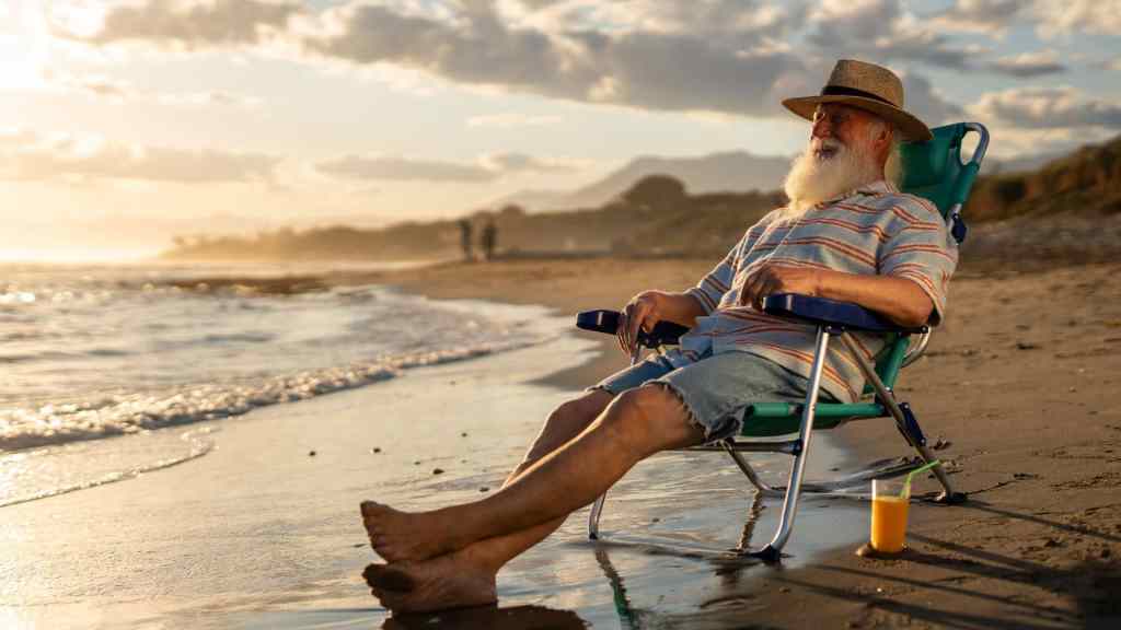 An older man with a white beard sits on a beach chair at sunset, wearing a hat and striped shirt, with a glass of orange juice on the sand beside him.