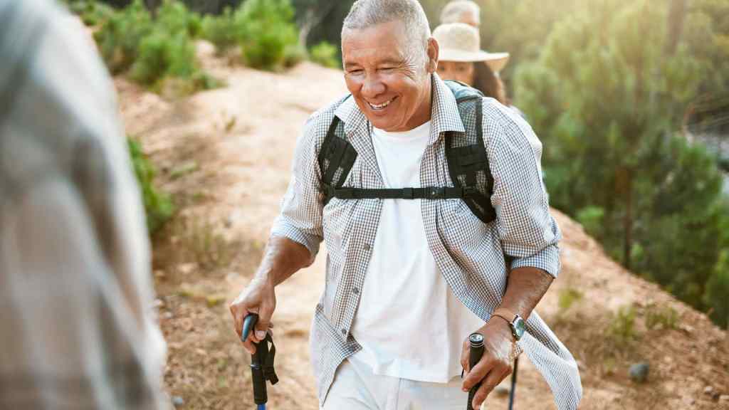 Older man hiking on a sunny trail, using trekking poles, wearing a black backpack and white shirt, with trees in the background.