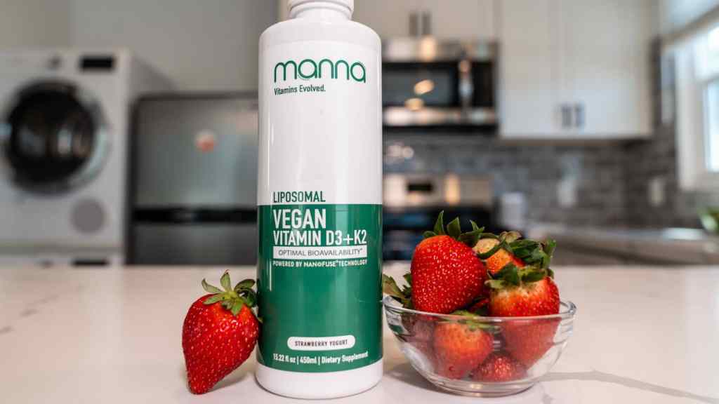 A bottle of Manna Liposomal Vegan Vitamin D3+K2 in strawberry flavor is on a kitchen counter next to a small glass bowl of fresh strawberries.