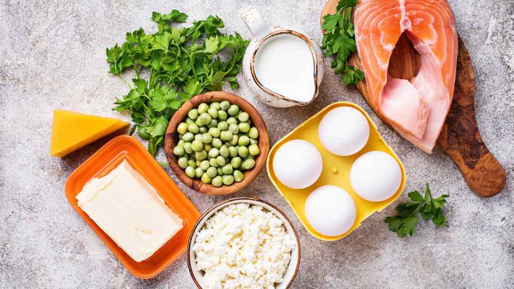 A selection of protein-rich foods on a table, including salmon, eggs, milk, cheese, cottage cheese, butter, green peas, and fresh parsley.