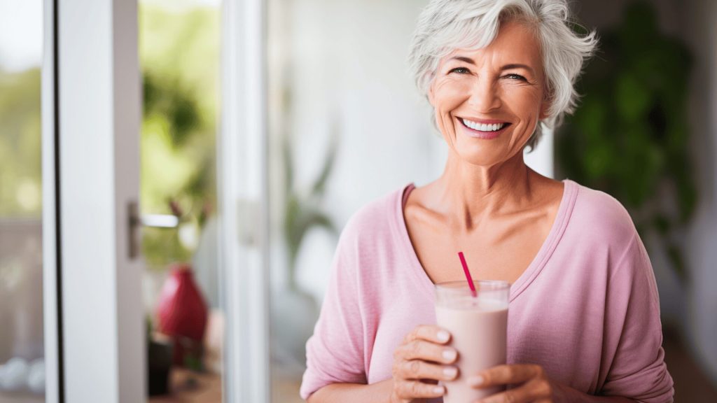 Smiling older woman with short gray hair holds a glass of a pink smoothie with a straw, standing indoors near a window.