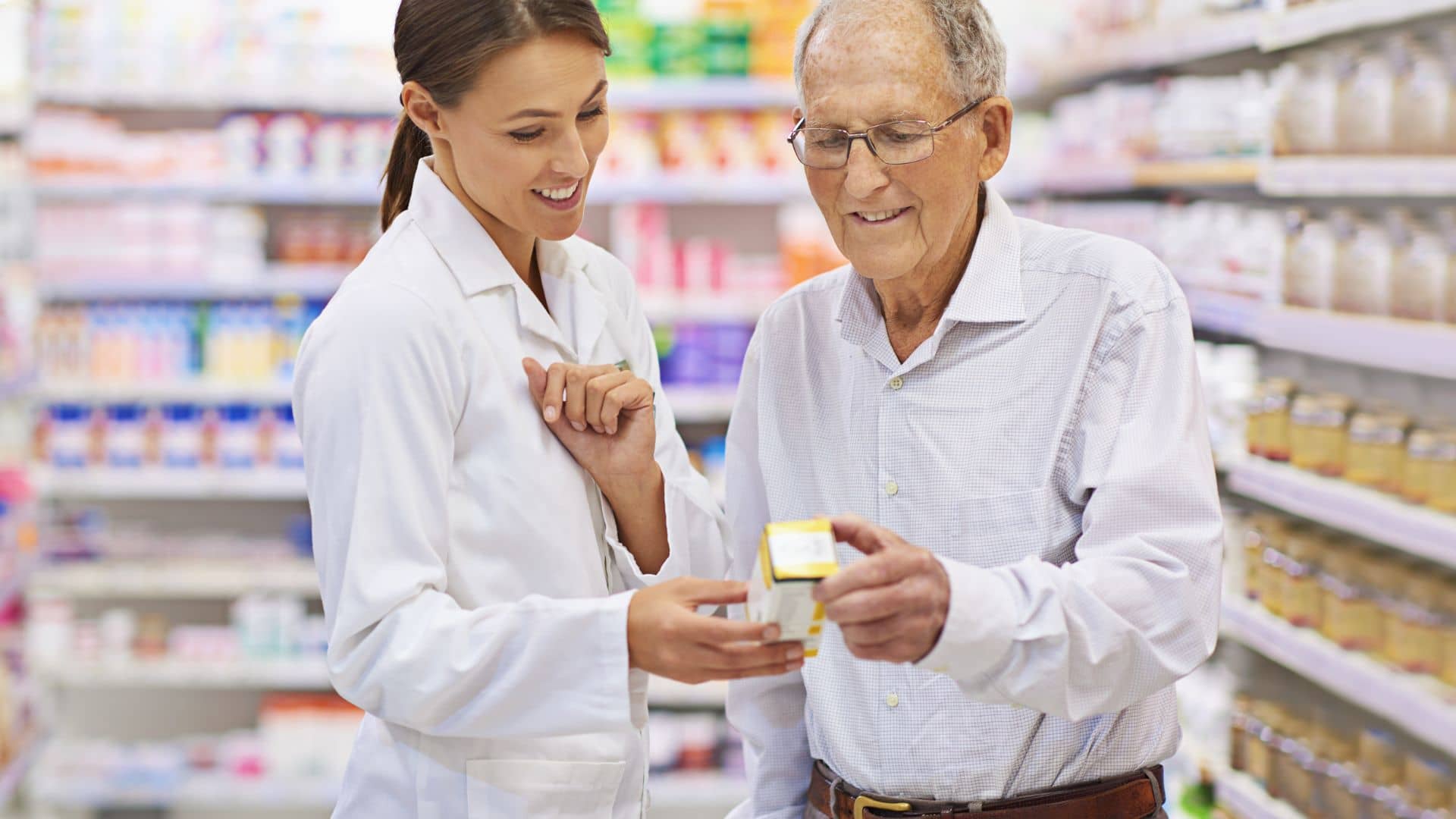 A pharmacist helps an elderly man choose a medication in a pharmacy aisle.