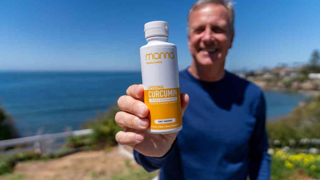 A person holding a bottle of liposomal curcumin supplement outdoors near a coastline on a sunny day.