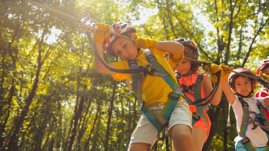 Children in helmets and harnesses navigate a rope course in a forested area on a sunny day.