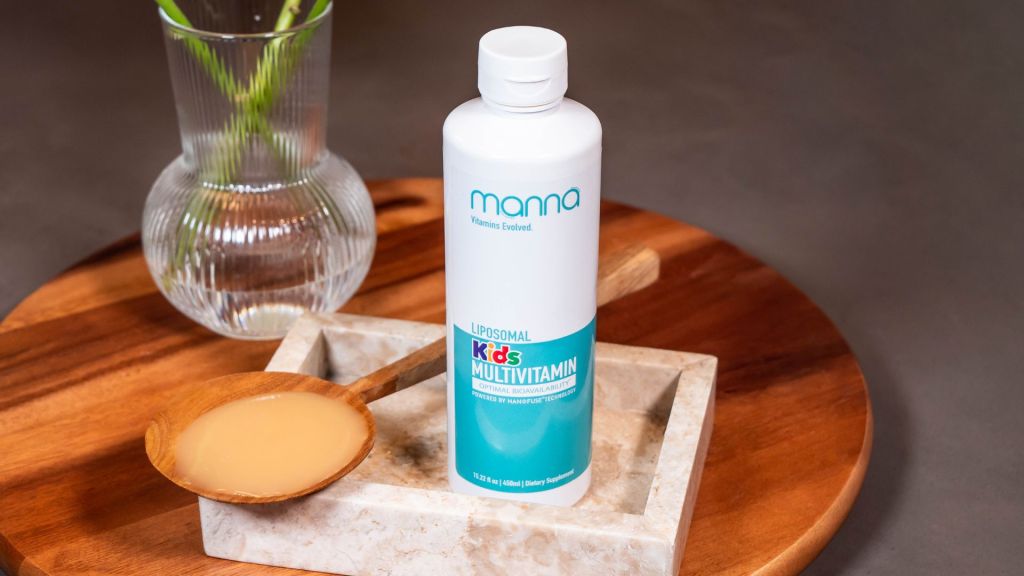 A bottle of Manna Liposomal Kids Multivitamin is placed on a wooden tray with a spoon containing liquid multivitamin, next to a vase with green stems.