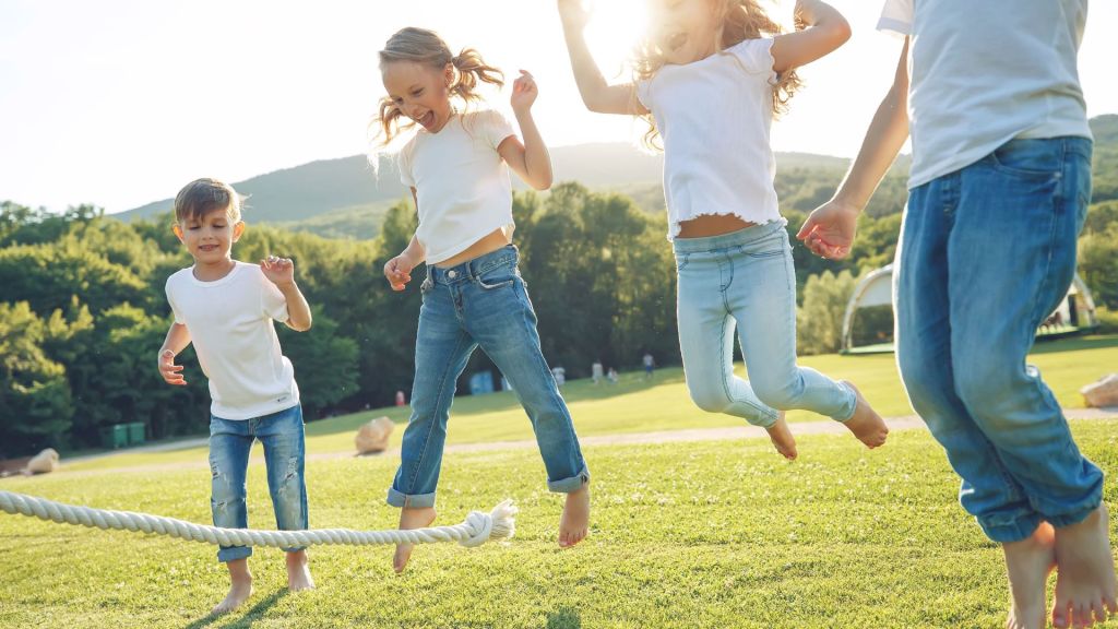 Children in jeans and white shirts are joyfully jumping over a rope on a sunny day in a grassy park.