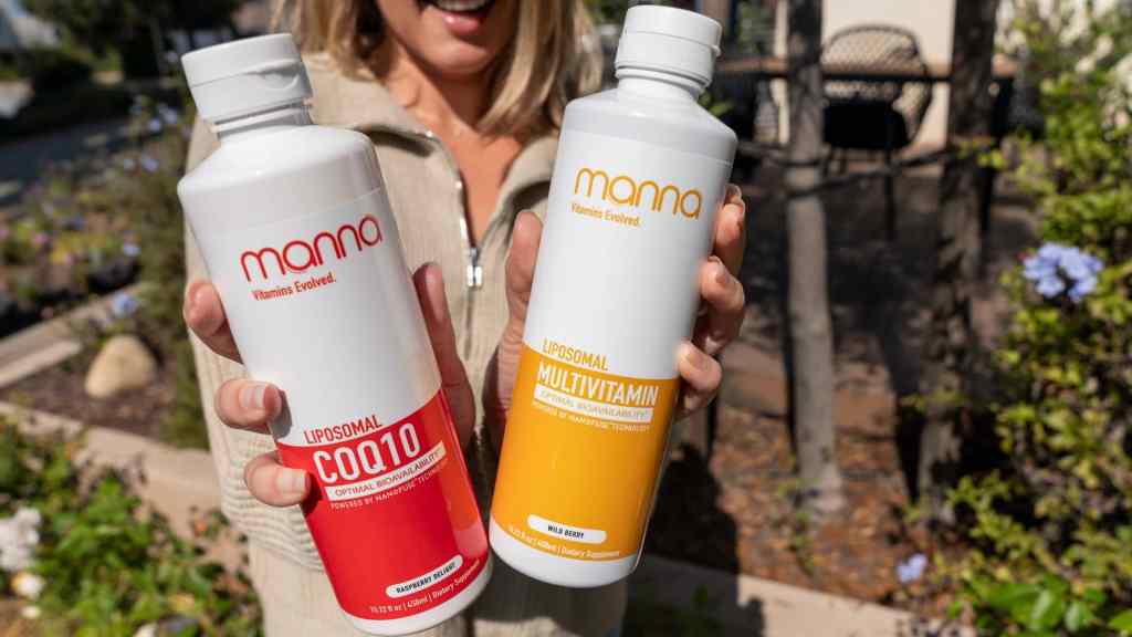 A person holding two bottles labeled "Manna Liposomal COQ10" and "Manna Liposomal Multivitamin" outdoors.