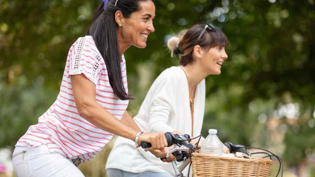 Two women riding bicycles outdoors, smiling. One has a striped shirt and the other is in white, with a wicker basket on the bike's front.