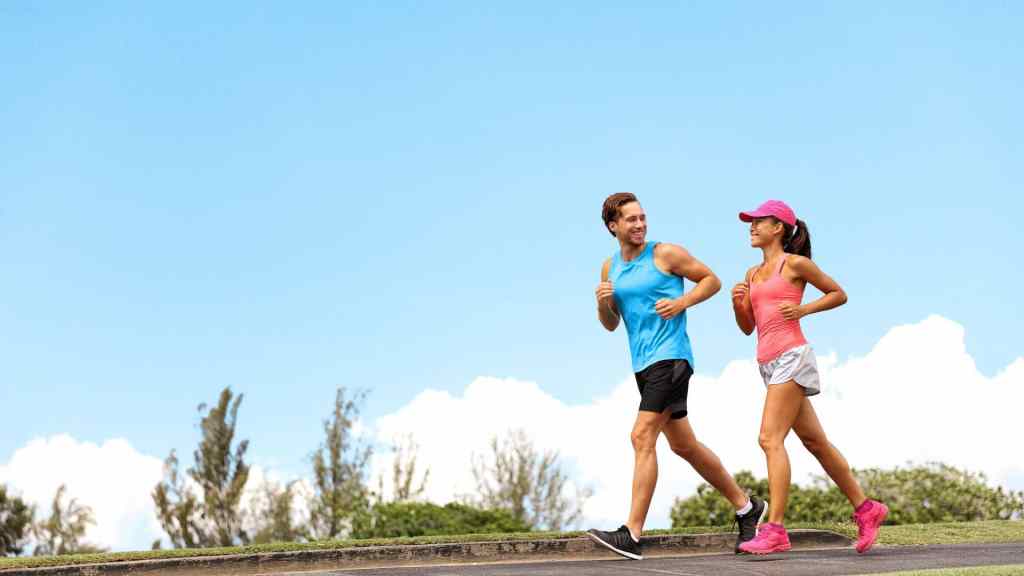A man and a woman are jogging outdoors on a clear day. The man is wearing a blue tank top and black shorts, while the woman has a pink tank top, white shorts, and a pink cap.