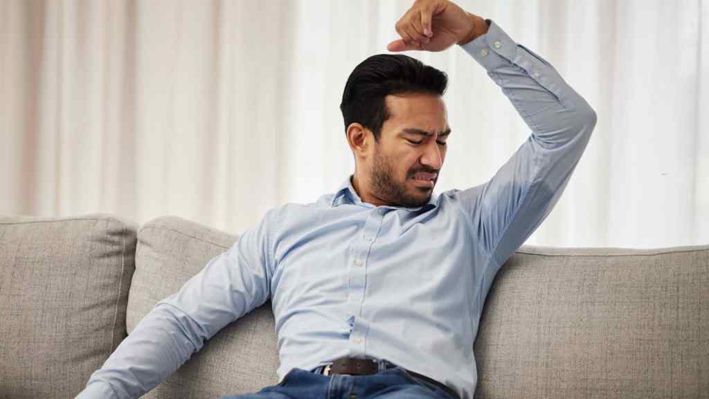 A man sitting on a couch lifts his arm and reacts in disgust while smelling his armpit.