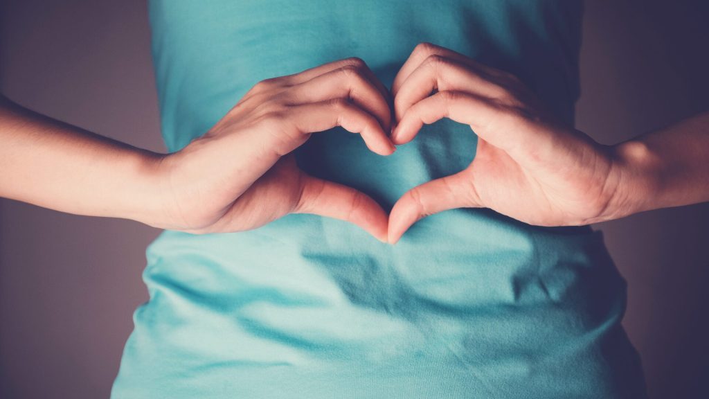 Close-up of hands forming a heart shape in front of a torso wearing a teal shirt.