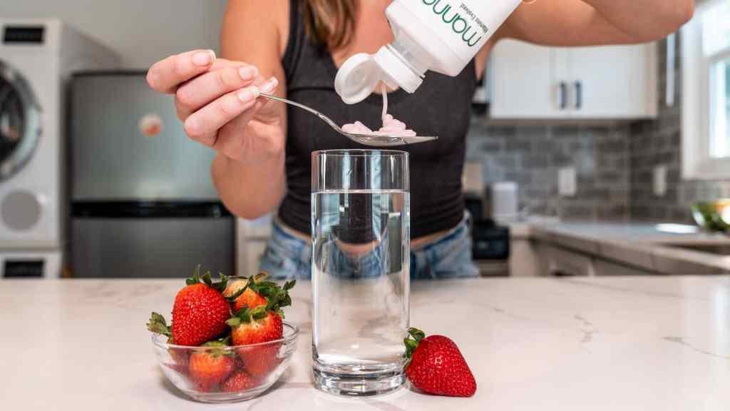 A person pours pink pills from a bottle onto a spoon over a glass of water, with a bowl of strawberries and a single strawberry on a kitchen counter.