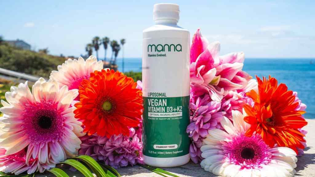 A bottle of Manna Liposomal Vegan Vitamin D3+K2 supplement stands among colorful flowers with the ocean and palm trees visible in the background.
