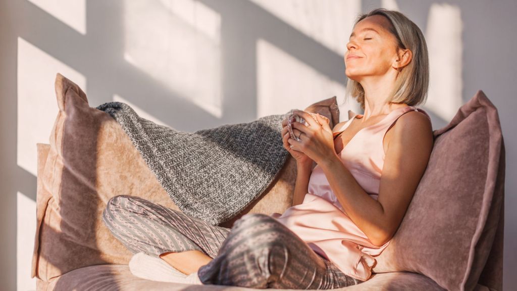 A woman sits comfortably on a plush couch, holding a cup and basking in the sunlight with her eyes closed, appearing relaxed.
