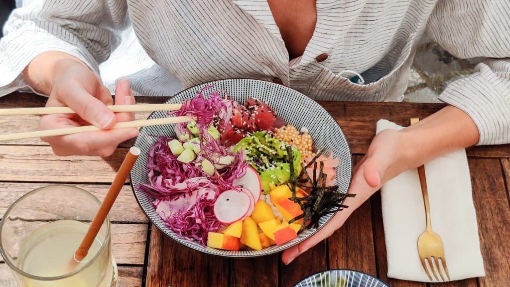A person holds a bowl of colorful poke with chopsticks. The bowl contains diced vegetables, fruits, seaweed, and raw fish. A glass of beverage, a cloth napkin, and a fork are on the wooden table.