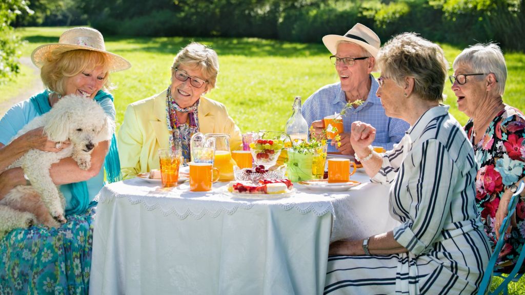 A group of seniors enjoy an outdoor gathering around a table with beverages and snacks. One person holds a small white dog. The setting is a sunny garden.