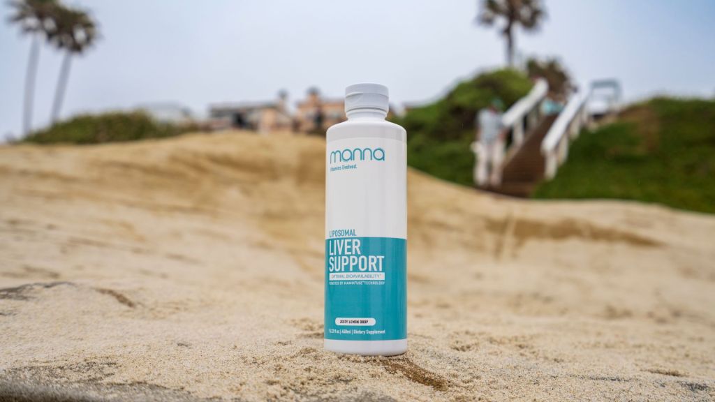 A bottle of "Personal Liver Support" dietary supplement is placed on a sandy beach with blurred greenery and steps in the background.