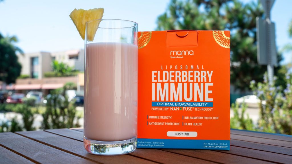A glass of light pink drink with a pineapple wedge on the rim sits next to a box of "Mama Liposomal Elderberry Immune" dietary supplement on a wooden table outdoors.