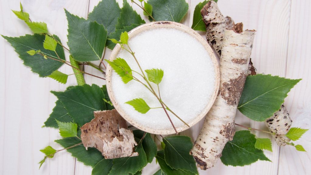 A bowl of white sugar sits on green leaves with birch bark and twigs surrounding it on a light wooden surface.
