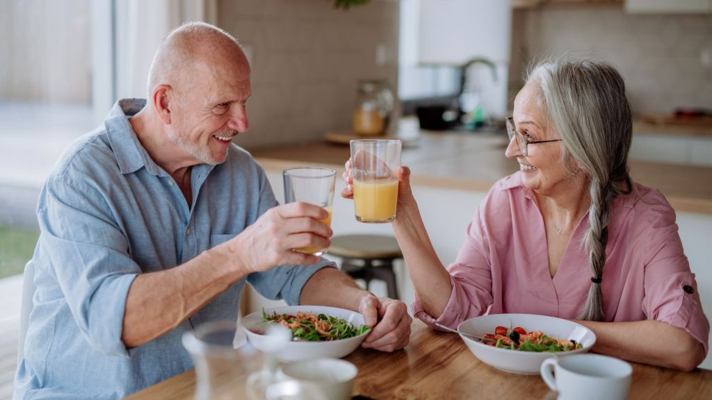 Two older adults sit at a dining table, smiling and toasting with glasses of juice. They have plates of food in front of them.