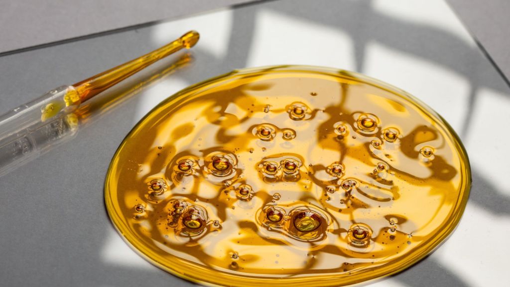A glass dropper beside a pool of viscous amber liquid on a reflective surface.