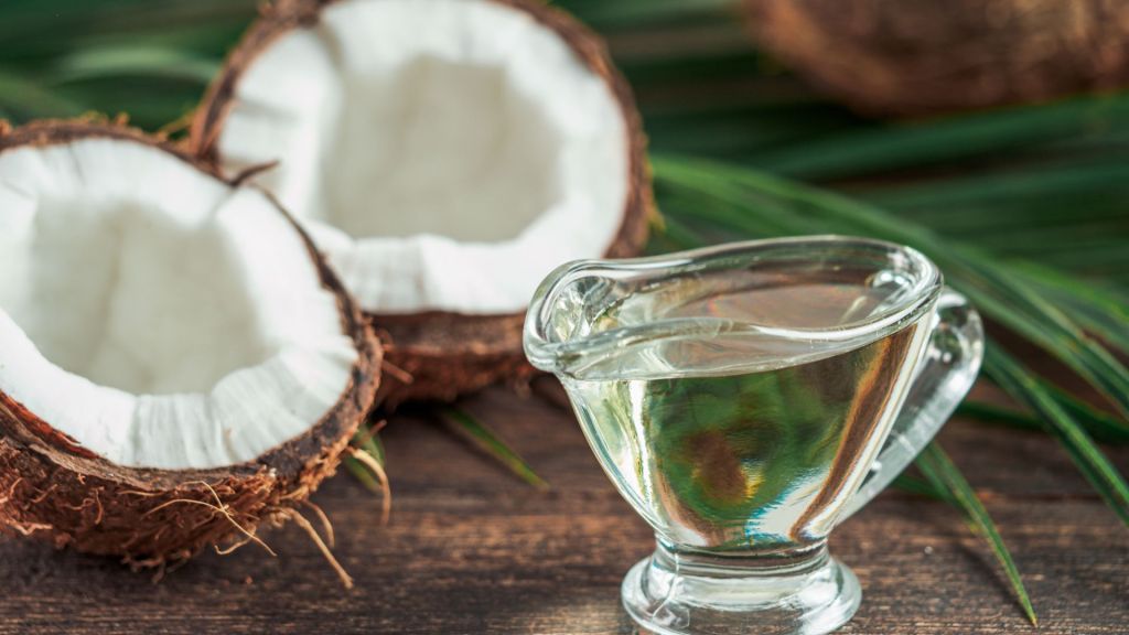 Glass pitcher of coconut oil next to halved coconuts on a wooden surface with green leaves in the background.