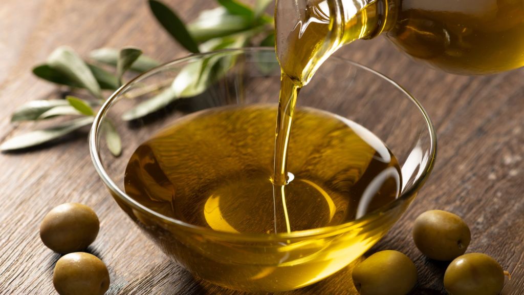 Olive oil being poured into a clear glass bowl, surrounded by green olives and olive leaves on a wooden surface.