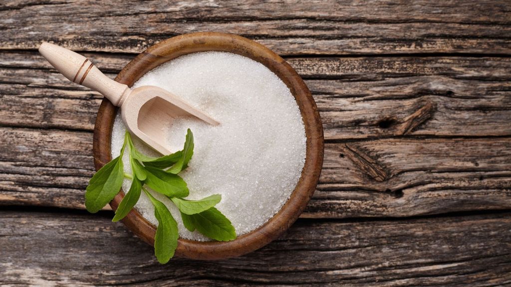 A wooden bowl of white sugar with a wooden scoop and green leaves on a wooden surface.