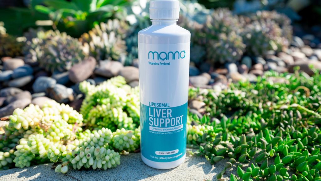 A bottle labeled "Manna Liposomal Liver Support" is placed on a concrete surface surrounded by green succulents and rocks.