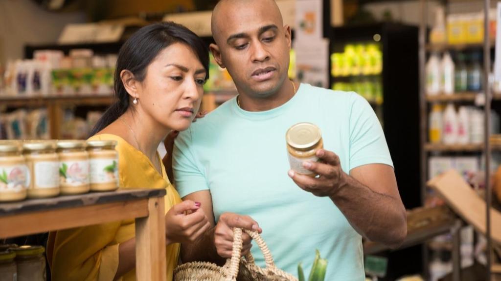 Man and woman holding a basket in a grocery store, looking at the back of a jar together.