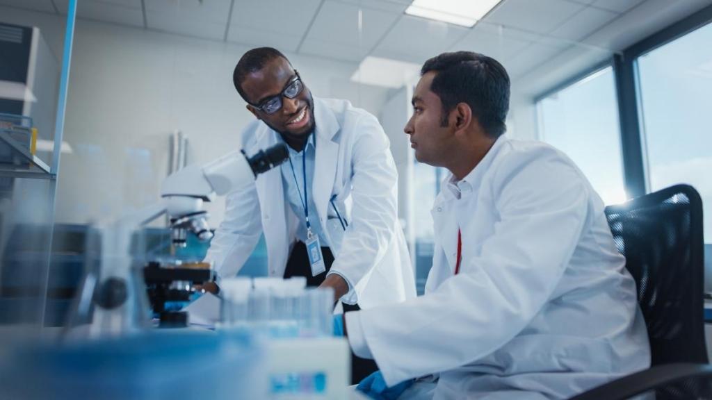 Two male scientists sitting and front of a microscope and various other lab supplies, talking.