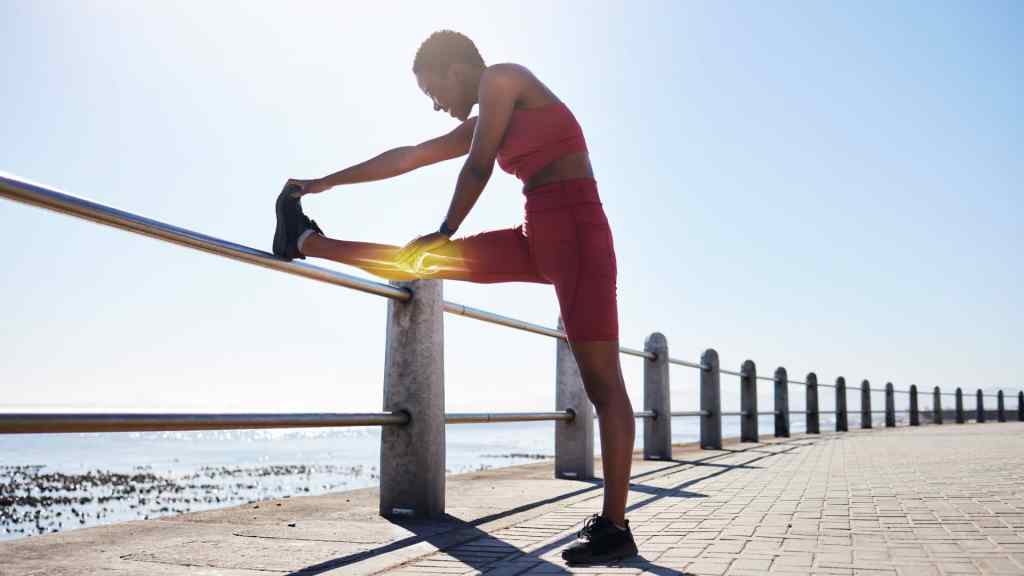 Person in red athletic wear stretching their leg on a railing by a waterside path in bright sunlight.