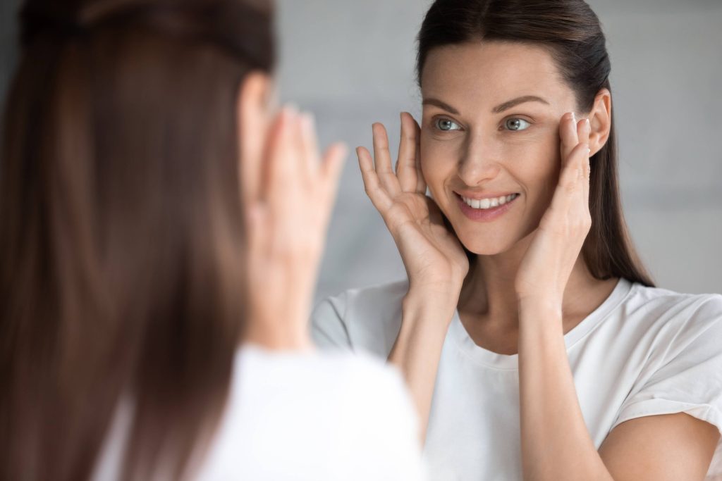A woman with long brown hair smiles as she looks at herself in the mirror, wearing a white shirt and touching her face with both hands.