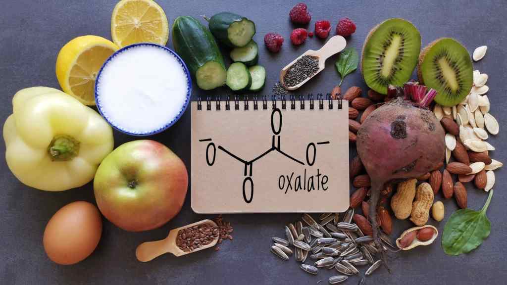 A notebook with an oxalate molecule diagram is surrounded by high-oxalate foods including fruits, vegetables, nuts, seeds, and eggs on a dark surface.