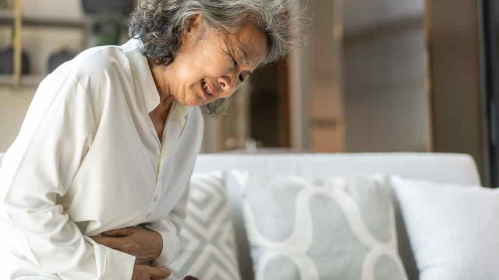 Older woman wearing white clothes sits on a couch, holding her stomach and appearing to be in pain or discomfort.
