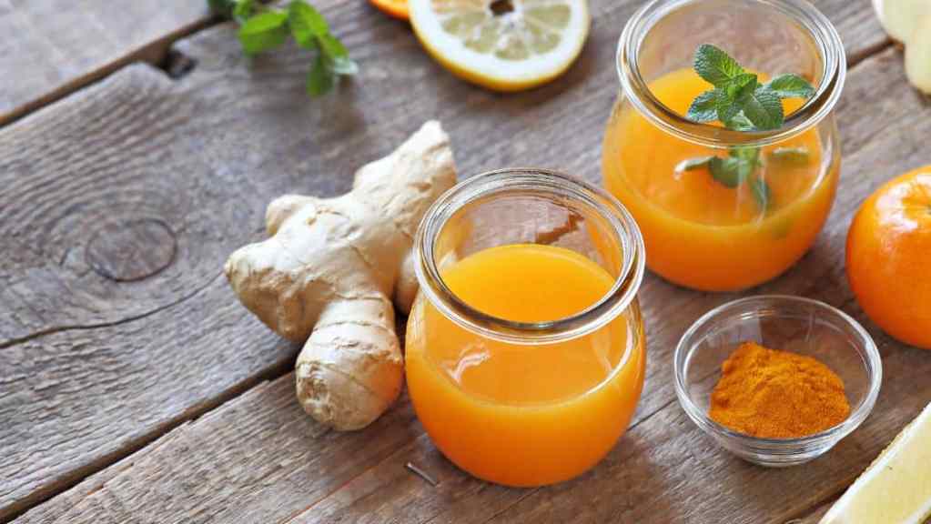 Two jars of fresh orange juice with mint leaves, surrounded by oranges, ginger, lemon, and turmeric on a wooden table.
