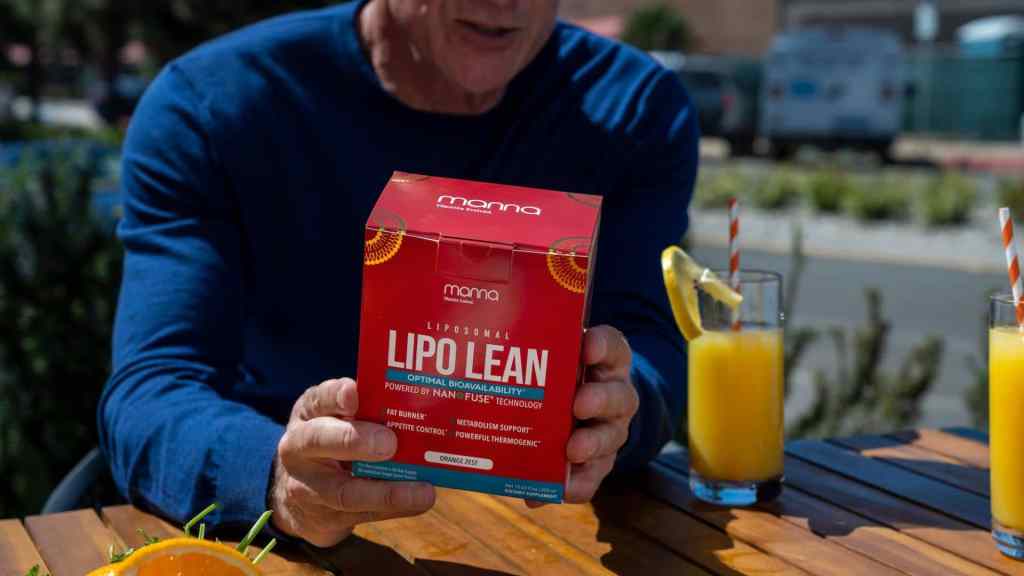 A person in a blue shirt is holding a red box labeled "Lipo Lean" at a wooden table with two glasses of juice, visible outdoor elements in the background.